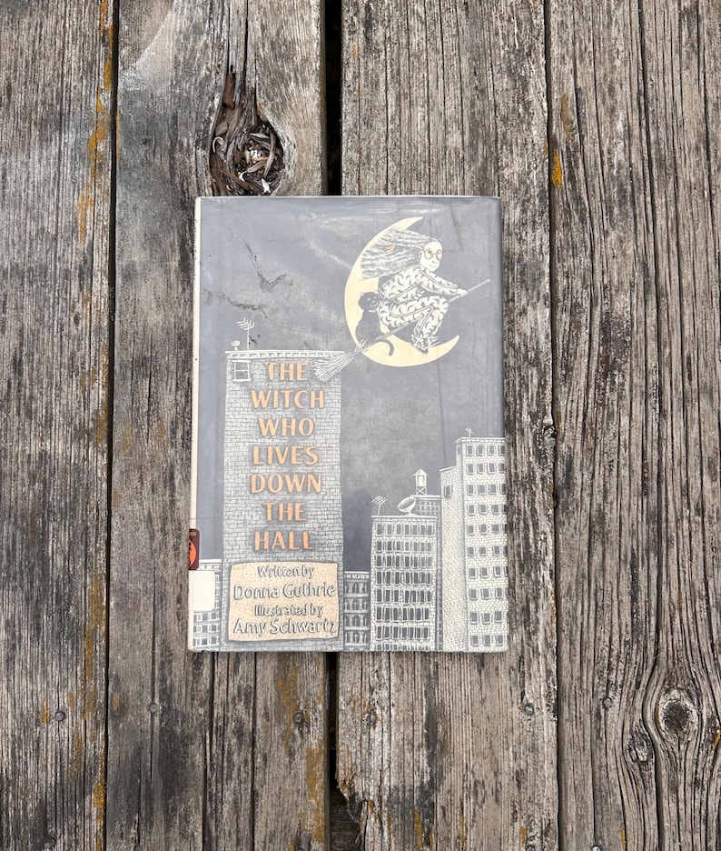 Puede incluir: Un libro de tapa dura titulado The Witch Who Lives Down the Hall, escrito por Donna Guthrie e ilustrado por Amy Schwartz. La portada presenta una ilustraci&oacute;n en blanco y negro de un paisaje urbano y una bruja en una escoba ante una luna amarilla.