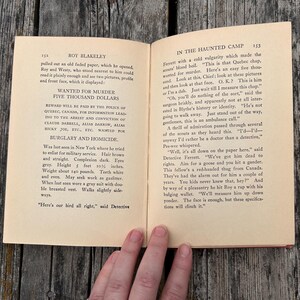 May include: An open vintage book titled Roy Blakeley In The Haunted Camp, pages 152 and 153. The left page features a printed wanted poster for murder with a five thousand dollar reward, while the right page contains narrative text in black ink.
