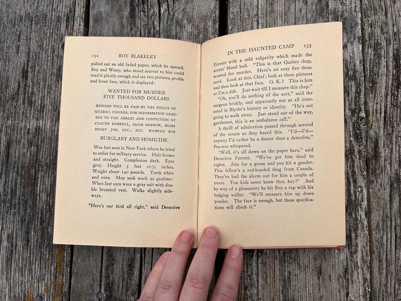 May include: An open vintage book titled Roy Blakeley In The Haunted Camp, pages 152 and 153. The left page features a printed wanted poster for murder with a five thousand dollar reward, while the right page contains narrative text in black ink.