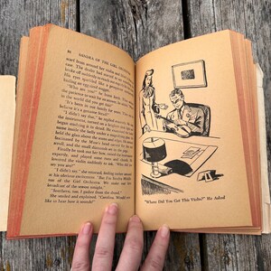 May include: An open vintage book with tan pages featuring black text and a black and white line art illustration of a person examining a violin. The text includes the title Sandra of the Girl Orchestra and the caption Where Did You Get This Violin? He Asked.