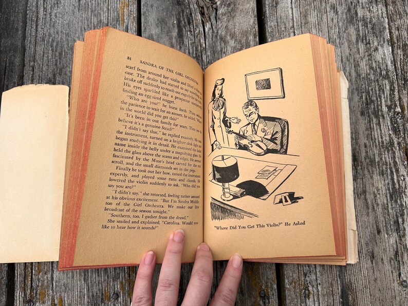 May include: An open vintage book with tan pages featuring black text and a black and white line art illustration of a person examining a violin. The text includes the title Sandra of the Girl Orchestra and the caption Where Did You Get This Violin? He Asked.