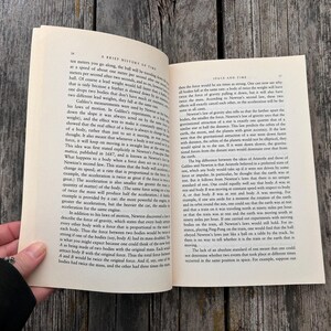 Peut inclure: Livre ouvert sur une surface en bois, affichant du texte. Le titre du livre est visible : "A Brief History of Time" et "Space and Time". Une main maintient le livre ouvert.