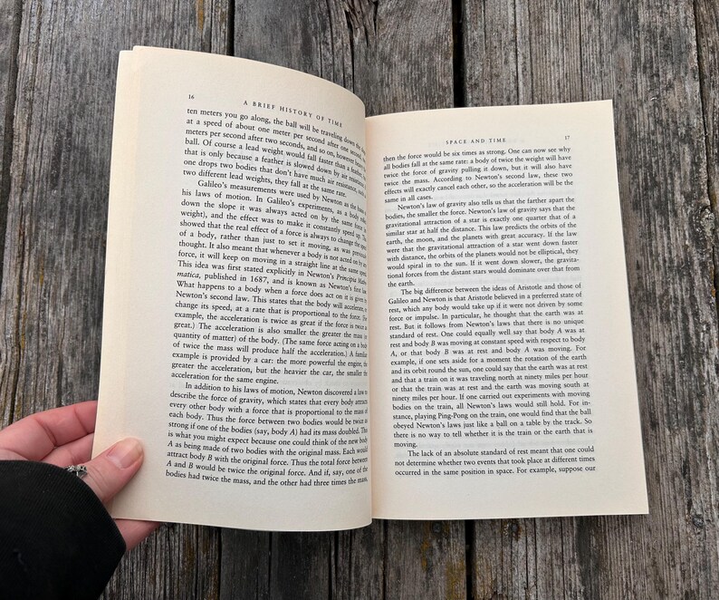 Peut inclure: Livre ouvert sur une surface en bois, affichant du texte. Le titre du livre est visible : "A Brief History of Time" et "Space and Time". Une main maintient le livre ouvert.