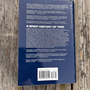 Peut inclure: Livre "Une br&egrave;ve histoire du temps" de Stephen Hawking. La couverture est bleu fonc&eacute; avec du texte blanc, avec des citations et un r&eacute;sum&eacute; du contenu du livre. Le livre est sur une surface en bois.