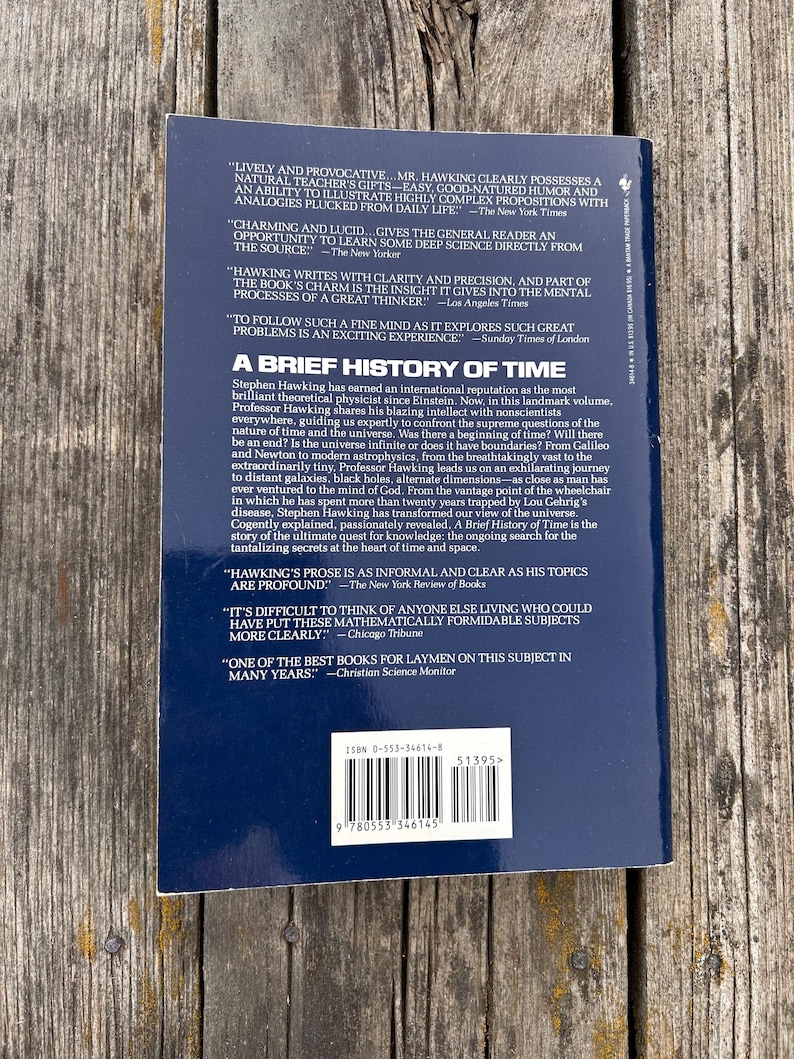 Peut inclure: Livre "Une br&egrave;ve histoire du temps" de Stephen Hawking. La couverture est bleu fonc&eacute; avec du texte blanc, avec des citations et un r&eacute;sum&eacute; du contenu du livre. Le livre est sur une surface en bois.