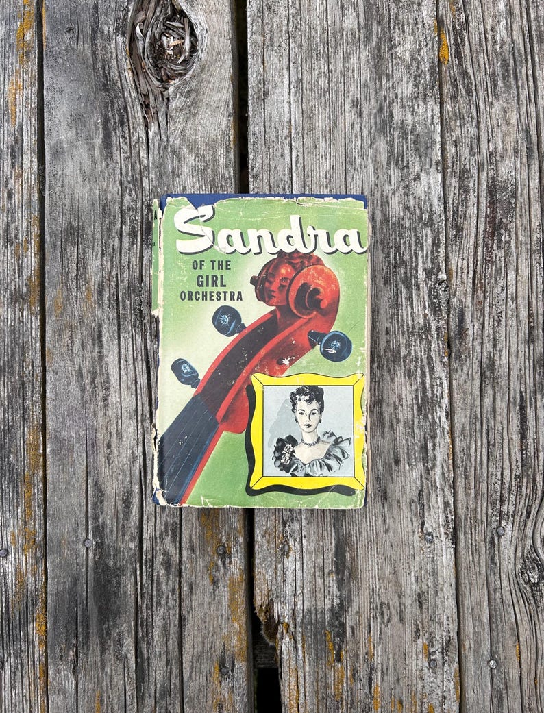 May include: A vintage hardback book titled Sandra of the Girl Orchestra with a green cover featuring a red violin scroll and a small framed illustration of a person. The book shows signs of wear along the edges and corners of the dust jacket.