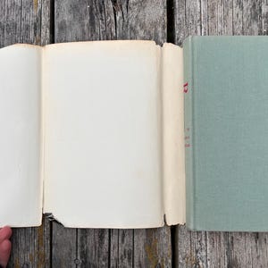 May include: A hardback book with a sage green fabric cover and a detached, cream-coloured paper dust jacket. The spine of the book shows a partial red letter R printed vertically. The pages are off-white and appear to be aged with light wear.