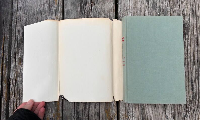 May include: A hardback book with a sage green fabric cover and a detached, cream-coloured paper dust jacket. The spine of the book shows a partial red letter R printed vertically. The pages are off-white and appear to be aged with light wear.