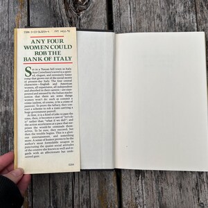 May include: A hardcover book with an open dust jacket flap featuring the title Any Four Women Could Rob the Bank of Italy in bold black text. The cream-colored flap contains a printed synopsis of the novel, while the book pages are white.