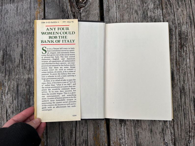 May include: A hardcover book with an open dust jacket flap featuring the title Any Four Women Could Rob the Bank of Italy in bold black text. The cream-colored flap contains a printed synopsis of the novel, while the book pages are white.
