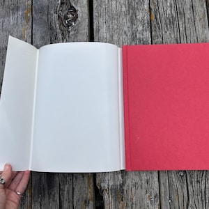 Op de afbeelding: Een hardcover boek met een felrode kaft en een uitgevouwen witte stofomslagflap. Het boek is geopend en toont lege witte pagina's, terwijl de kaft een effen, matte rode textuur heeft. De rug van het boek is recht en duidelijk gedefinieerd.
