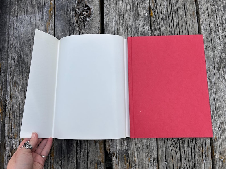 Op de afbeelding: Een hardcover boek met een felrode kaft en een uitgevouwen witte stofomslagflap. Het boek is geopend en toont lege witte pagina's, terwijl de kaft een effen, matte rode textuur heeft. De rug van het boek is recht en duidelijk gedefinieerd.