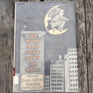 Puede incluir: Un libro de tapa dura titulado The Witch Who Lives Down the Hall, escrito por Donna Guthrie e ilustrado por Amy Schwartz. La portada muestra un horizonte urbano gris con una bruja volando en una escoba frente a una gran luna creciente.