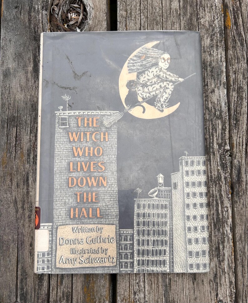 Puede incluir: Un libro de tapa dura titulado The Witch Who Lives Down the Hall, escrito por Donna Guthrie e ilustrado por Amy Schwartz. La portada muestra un horizonte urbano gris con una bruja volando en una escoba frente a una gran luna creciente.