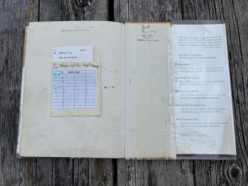 May include: An open vintage library book with a cream-colored interior. The left page features a paper checkout card pocket with a date due slip. The right page contains a printed summary of the Billy and Blaze book series with clear tape.