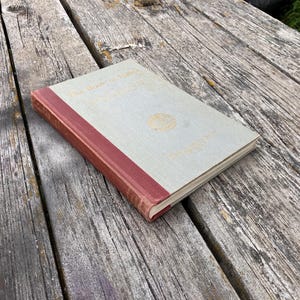 Può includere: Un libro d'epoca intitolato "The Book of Hours" con una copertina grigio chiaro e un dorso rosso. Il libro ha scritte dorate e un emblema dorato sulla copertina. Il libro è appoggiato su una superficie di legno usurata.