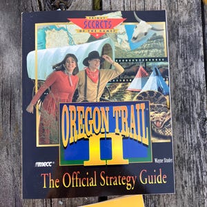 Puede incluir: Portada de la guía de estrategia del juego "Oregon Trail II". Muestra una pareja con ropa de época, una carreta cubierta, tipis y una serpiente de cascabel. El título está en letras grandes amarillas y azules.