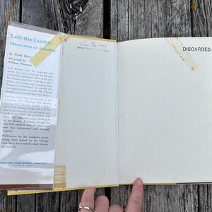 Pu&ograve; includere: Un libro con copertina rigida aperto intitolato Leif the Lucky: Discoverer of America di Erick Berry. La copertina interna presenta un riassunto incollato con nastro adesivo ingiallito, mentre la pagina opposta &egrave; vuota con la scritta DISCARDED.