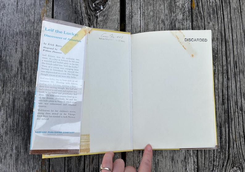 Pu&ograve; includere: Un libro con copertina rigida aperto intitolato Leif the Lucky: Discoverer of America di Erick Berry. La copertina interna presenta un riassunto incollato con nastro adesivo ingiallito, mentre la pagina opposta &egrave; vuota con la scritta DISCARDED.