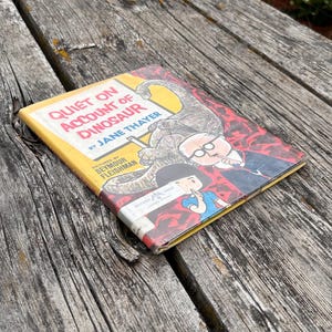 Pode incluir: Um livro infantil de capa dura intitulado Quiet on Account of Dinosaur de Jane Thayer, com ilustra&ccedil;&otilde;es de Seymour Fleishman. A capa apresenta um grande dinossauro, uma pessoa de &oacute;culos e uma crian&ccedil;a fazendo um gesto de sil&ecirc;ncio.