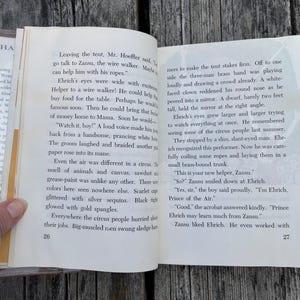 May include: An open book with white pages and black printed text, showing pages 26 and 27. The left page begins with the text Leaving the tent, Mr. Hoeffler said, and the right page ends with the text Zansu liked Ehrich. He even worked with.