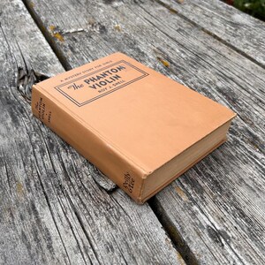 K&ouml;nnte beinhalten: Ein beigefarbenes gebundenes Buch mit dem Titel The Phantom Violin von Roy J. Snell. Das Cover zeigt schwarzen Text in einem rechteckigen Rahmen, wobei Titel und Autor auch auf dem Buchr&uuml;cken stehen. Der Verlag Reilly & Lee steht auf dem R&uuml;cken.