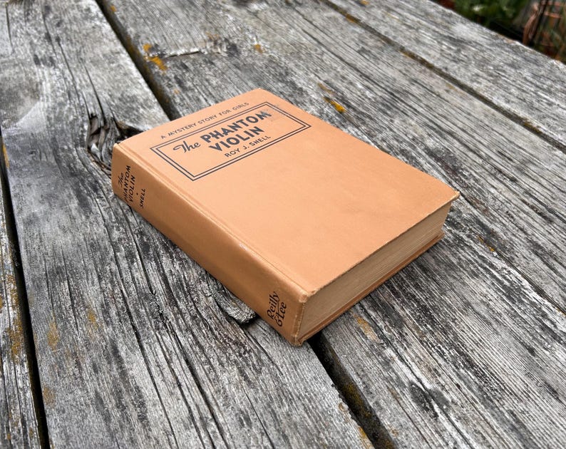 K&ouml;nnte beinhalten: Ein beigefarbenes gebundenes Buch mit dem Titel The Phantom Violin von Roy J. Snell. Das Cover zeigt schwarzen Text in einem rechteckigen Rahmen, wobei Titel und Autor auch auf dem Buchr&uuml;cken stehen. Der Verlag Reilly & Lee steht auf dem R&uuml;cken.