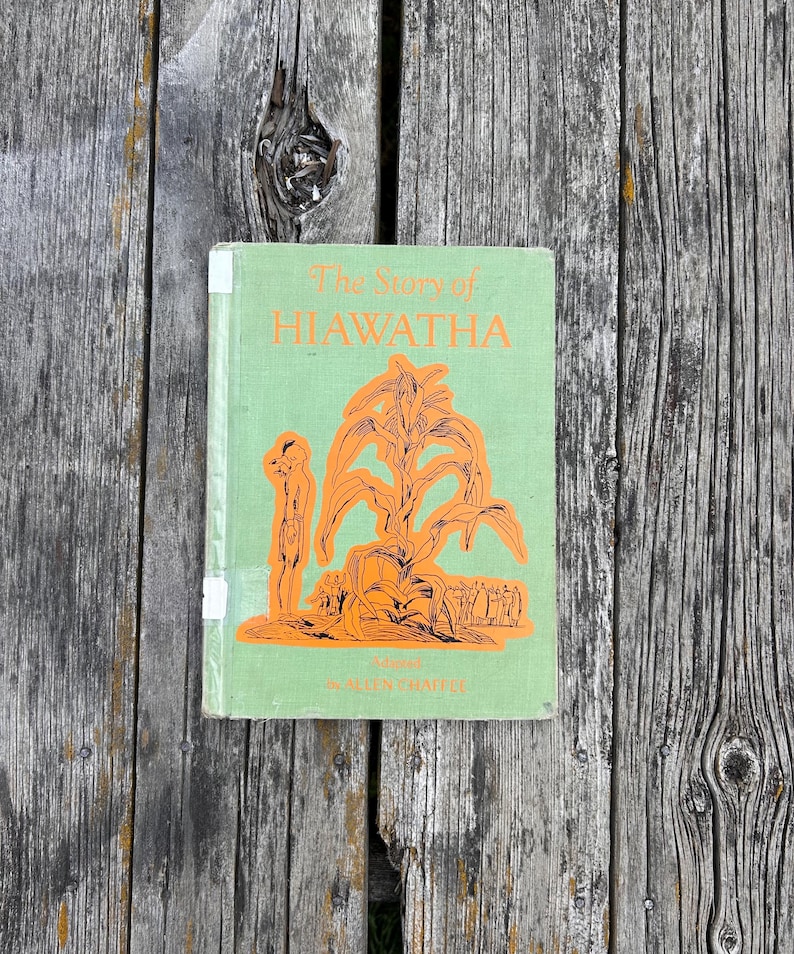 K&ouml;nnte beinhalten: Ein gebundenes Buch mit dem Titel The Story of HIAWATHA, adaptiert von Allen Chappel. Der Einband ist hellgr&uuml;n mit orangefarbenen Illustrationen einer Person neben einem gro&szlig;en Maiskolben und einer Menschengruppe. Der Titel ist orange.