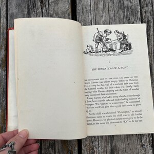 May include: An open hardcover book with cream-colored pages featuring black text and a line art illustration of two figures near a crib. The chapter title reads I THE EDUCATION OF A RUNT, followed by printed paragraphs of narrative text.