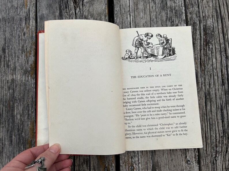May include: An open hardcover book with cream-colored pages featuring black text and a line art illustration of two figures near a crib. The chapter title reads I THE EDUCATION OF A RUNT, followed by printed paragraphs of narrative text.