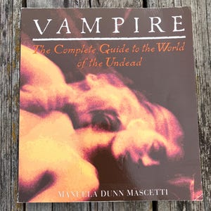 May include: A book titled "VAMPIRE" with the subtitle "The Complete Guide to the World of the Undead." The cover features an abstract image in shades of orange and brown. The author's name, MANUELA DUNN MASCETTI, is printed at the bottom.