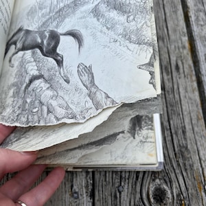 May include: A book with pages featuring black and white pencil illustrations of a horse jumping over a stone wall and a person with hands raised. The text on the page reads, "top him, he and jumped a high oss the fields."