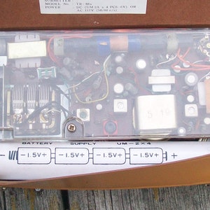 Może przedstawiać: Przenośne radio vintage z otwartym brązowym sk&oacute;rzanym etui, ukazujące wewnętrzne komponenty elektroniczne pod przezroczystą plastikową pokrywą. Biała etykieta komory baterii zawiera schemat dla czterech baterii 1,5V, z napisem BATTERY SUPPLY UM-2x4.