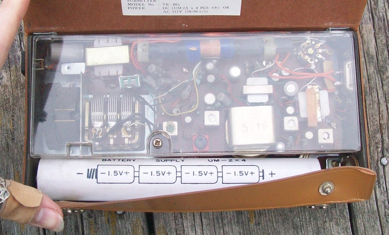 Może przedstawiać: Przenośne radio vintage z otwartym brązowym sk&oacute;rzanym etui, ukazujące wewnętrzne komponenty elektroniczne pod przezroczystą plastikową pokrywą. Biała etykieta komory baterii zawiera schemat dla czterech baterii 1,5V, z napisem BATTERY SUPPLY UM-2x4.