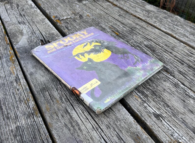 K&ouml;nnte beinhalten: Ein gebundenes Kinderbuch mit dem Titel Spooky and the Bad Luck Raven von Natalie Savage Carlson. Der violette Einband zeigt einen gelben Mond, eine schwarze Katze und einen Raben. Ein gelber Aufkleber der Bibliotheksausgabe ist zu sehen.