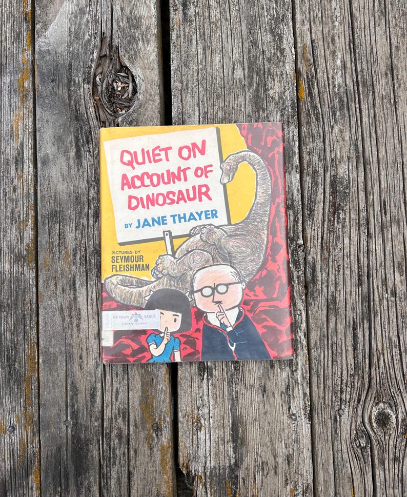 Pode incluir: Um livro infantil de capa dura intitulado Quiet on Account of Dinosaur de Jane Thayer com ilustra&ccedil;&otilde;es de Seymour Fleishman. A capa mostra um grande dinossauro castanho, uma rapariga e um homem de &oacute;culos a fazer um gesto de sil&ecirc;ncio.