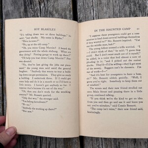 May include: An open vintage book titled Roy Blakeley with pages 20 and 21 visible. The text In The Haunted Camp is printed at the top of the right page. The paper is off-white with black printed text featuring dialogue between characters.