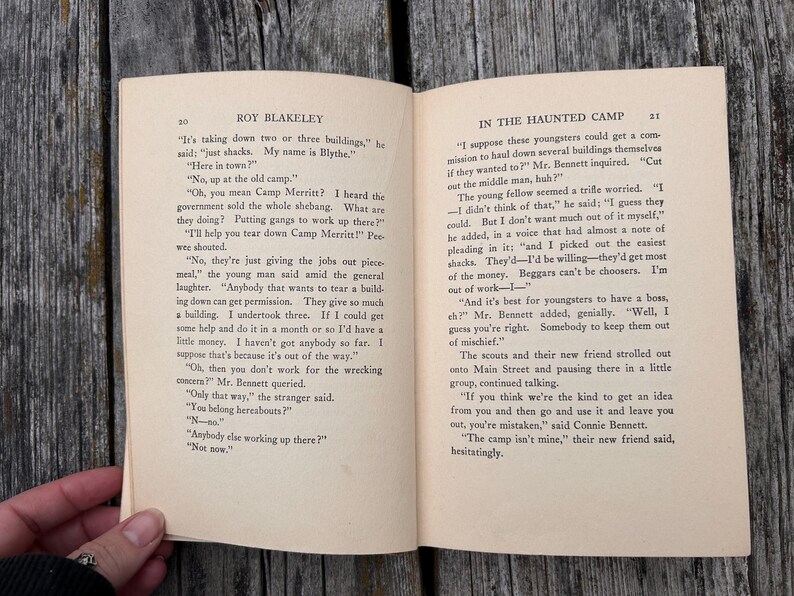 May include: An open vintage book titled Roy Blakeley with pages 20 and 21 visible. The text In The Haunted Camp is printed at the top of the right page. The paper is off-white with black printed text featuring dialogue between characters.