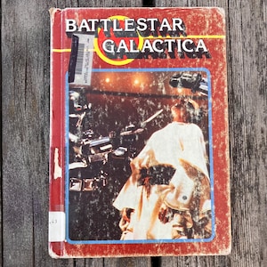 Op de afbeelding: Een vintage exemplaar van "Battlestar Galactica" met een rode omslag en de titel in vette, gele en witte letters. De omslag toont een scène uit de serie met een figuur in een witte jurk. Het boek is versleten.