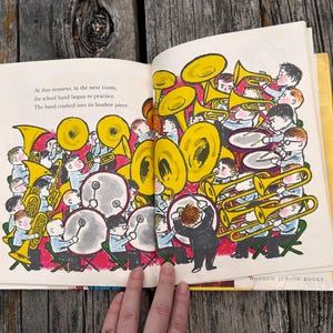 Pode incluir: Uma p&aacute;gina aberta de um livro infantil com uma ilustra&ccedil;&atilde;o colorida de uma banda escolar tocando instrumentos, incluindo trompas amarelas e tambores. O texto diz: At that moment, in the next room, the school band began to practice. The band crashed into its loudest piece.