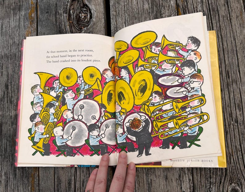 Pode incluir: Uma p&aacute;gina aberta de um livro infantil com uma ilustra&ccedil;&atilde;o colorida de uma banda escolar tocando instrumentos, incluindo trompas amarelas e tambores. O texto diz: At that moment, in the next room, the school band began to practice. The band crashed into its loudest piece.