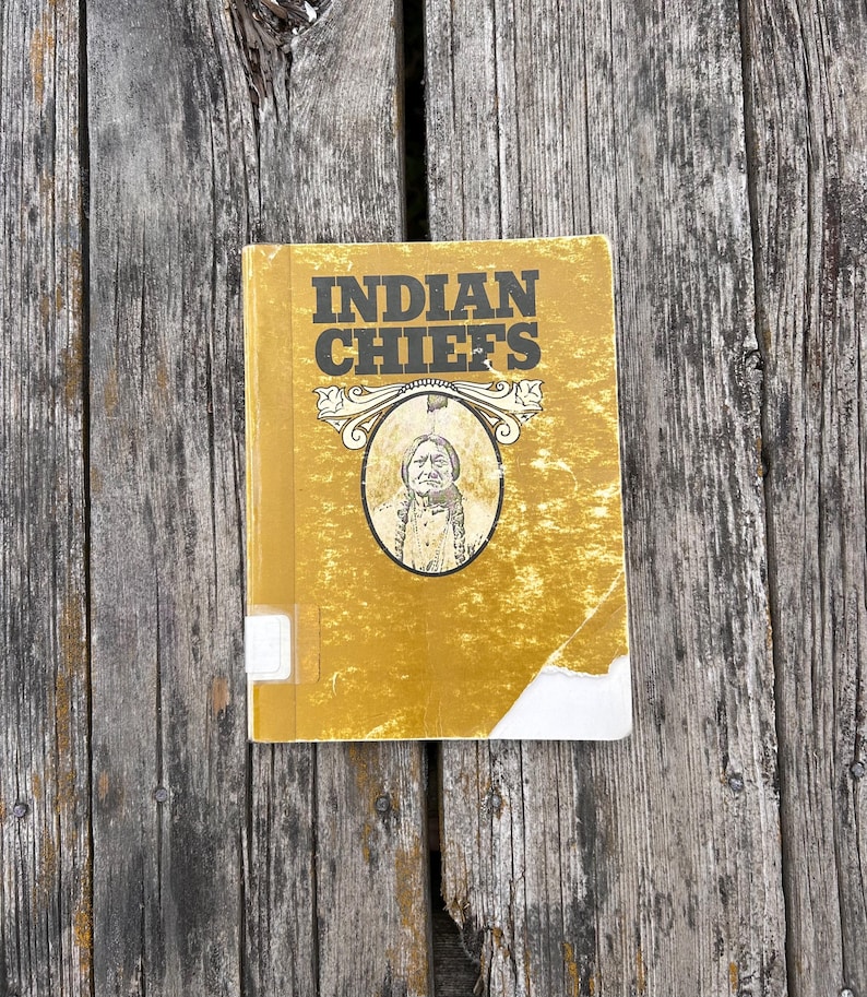 Może przedstawiać: Ż&oacute;łta książka w twardej oprawie z tytułem INDIAN CHIEFS pogrubionymi czarnymi literami. Okładka zawiera centralną owalną ilustrację osoby z warkoczami, ozdobne zawijasy nad portretem oraz małą białą etykietę na grzbiecie książki.