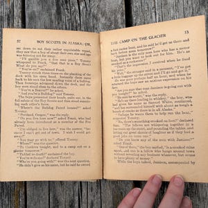 K&ouml;nnte beinhalten: Ein aufgeschlagenes altes Buch mit dem Titel Boy Scouts in Alaska: Or, The Camp on the Glacier, das zwei Seiten mit schwarzem Drucktext auf gealtertem, hellbraunem Papier zeigt. Die Seiten 52 und 53 enthalten einen dialogreichen Text.