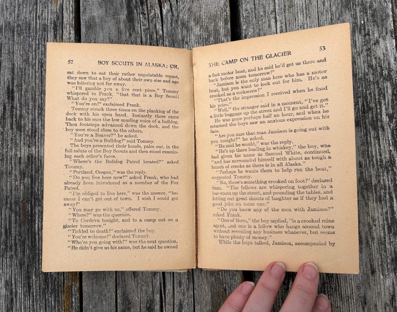 K&ouml;nnte beinhalten: Ein aufgeschlagenes altes Buch mit dem Titel Boy Scouts in Alaska: Or, The Camp on the Glacier, das zwei Seiten mit schwarzem Drucktext auf gealtertem, hellbraunem Papier zeigt. Die Seiten 52 und 53 enthalten einen dialogreichen Text.