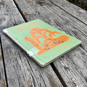 K&ouml;nnte beinhalten: Ein gebundenes Buch mit dem Titel The Song of Hiawatha und einem hellgr&uuml;nen Einband. Die Vorderseite zeigt eine orangefarbene Illustration einer Figur und stilisierter B&auml;ume. Der Buchr&uuml;cken ist mit wei&szlig;em Klebeband verst&auml;rkt.