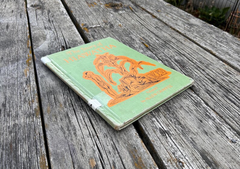 K&ouml;nnte beinhalten: Ein gebundenes Buch mit dem Titel The Song of Hiawatha und einem hellgr&uuml;nen Einband. Die Vorderseite zeigt eine orangefarbene Illustration einer Figur und stilisierter B&auml;ume. Der Buchr&uuml;cken ist mit wei&szlig;em Klebeband verst&auml;rkt.