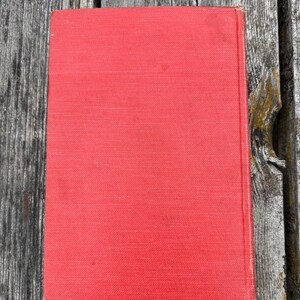 May include: A rectangular hardcover book with a solid, vibrant red fabric cover. The book features a clean, plain front surface with a visible vertical spine edge on the right side, showcasing a simple, uniform texture across the entire front cover.