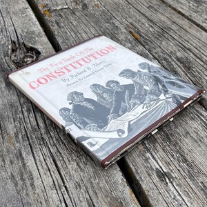 Puede incluir: Un libro de tapa dura titulado The First Book of The Constitution por Richard B. Morris con im&aacute;genes de Leonard Everett Fisher. La cubierta blanca presenta una ilustraci&oacute;n en blanco y negro de figuras hist&oacute;ricas firmando un documento.
