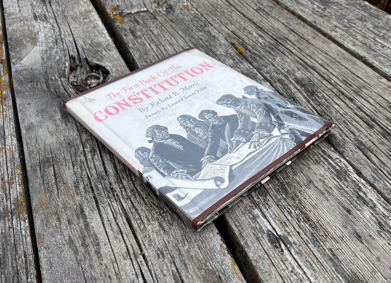 Puede incluir: Un libro de tapa dura titulado The First Book of The Constitution por Richard B. Morris con im&aacute;genes de Leonard Everett Fisher. La cubierta blanca presenta una ilustraci&oacute;n en blanco y negro de figuras hist&oacute;ricas firmando un documento.