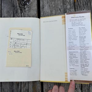 Pu&ograve; includere: Un libro con copertina rigida aperto con una scheda di prestito bibliotecario color crema in una tasca sulla pagina sinistra. La pagina destra presenta un elenco stampato intitolato Discovery Books, che dettaglia varie figure storiche.